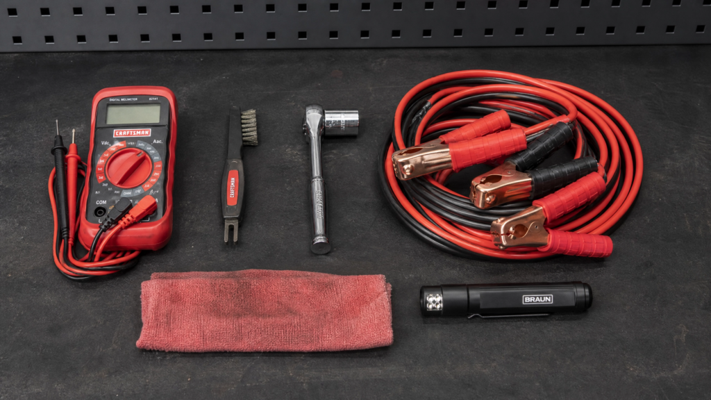 image of a mechanic's workbench with tools laid out neatly: a red digital multimeter, terminal cleaning brush, 10mm socket wrench, jumper cables coiled up, a shop rag, and a small LED flashlight